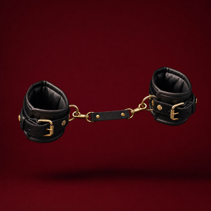 Bijou padded restraint cuff pair in black with gold buckles and a clip-on connector strap