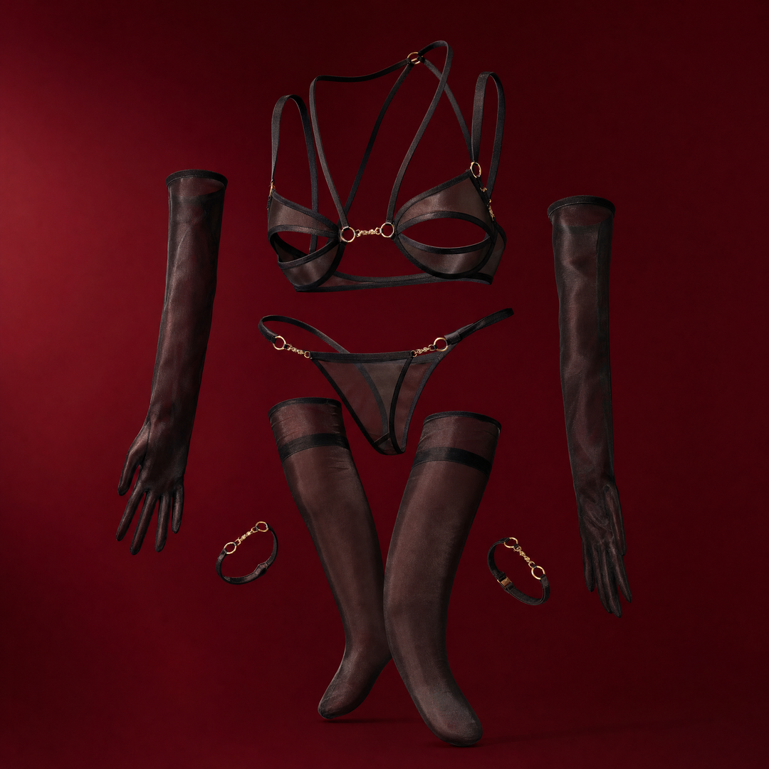 FLORA | CHAIN BRA SET - sheer black mesh set with gold hardware: open cut-out bra with gold script charm, matching thong, a pair of long sheer gloves, stockings and two charm leg bands, suspended on a deep burgundy studio background